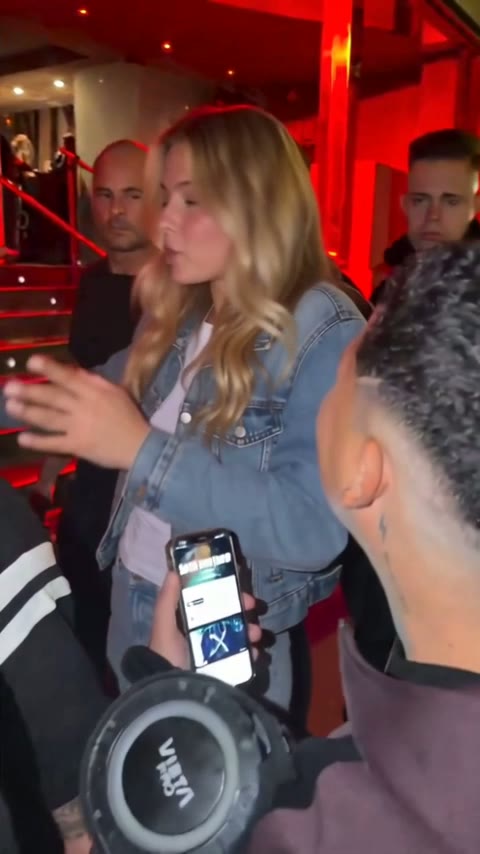Blonde woman with long waves and a denim jacket facing a phone held up in the night crowd
